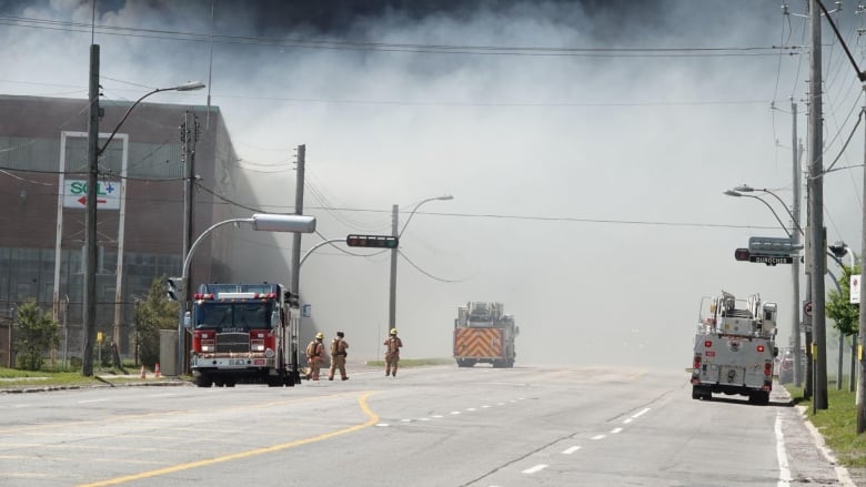 Three firefighters walk between fire trucks parked in front of an industrial building.
