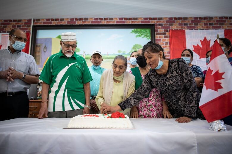 A group of Asian seniors stand around a cake. A woman in a yellow dress in the centre attempts to cut a cake, assisted by a woman with a mask around her chin.