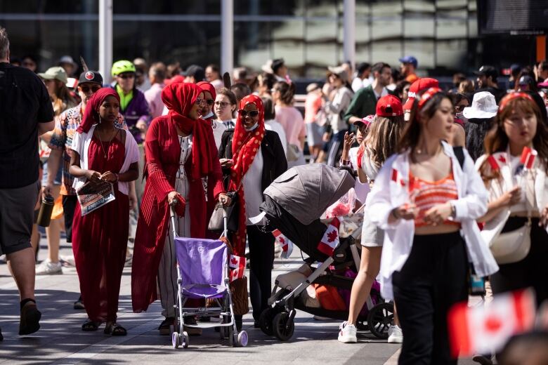 A large group of people, most of whom are wearing red to commemorate Canada Day. In focus are a family of three hijabi women. They all have red hijabs, and one of them is pushing a purple pushcart.