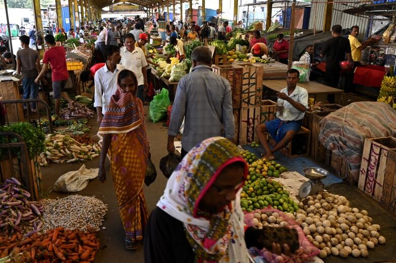 Gente caminando en un mercado de verduras con montones de frutas en verduras en el suelo y en las mesas. 