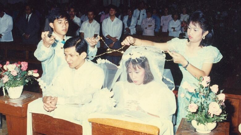 A Filipino couple kneels at wedding ceremony while a golden cord is held above their heads by another man and woman.