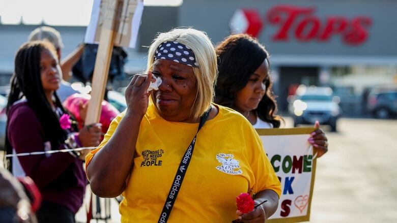 A woman wipes tears from her eyes outside a supermarket. Another woman behind her holds a sign that says "Welcome back." 