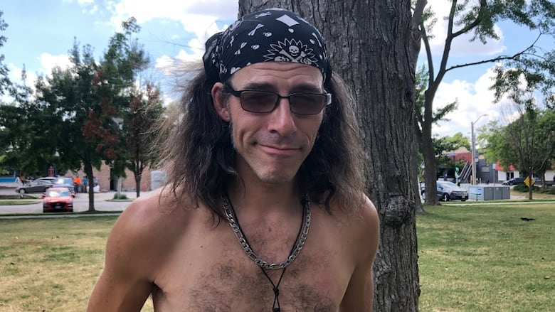 A shirtless man with sunglasses stands in front of a tree. 