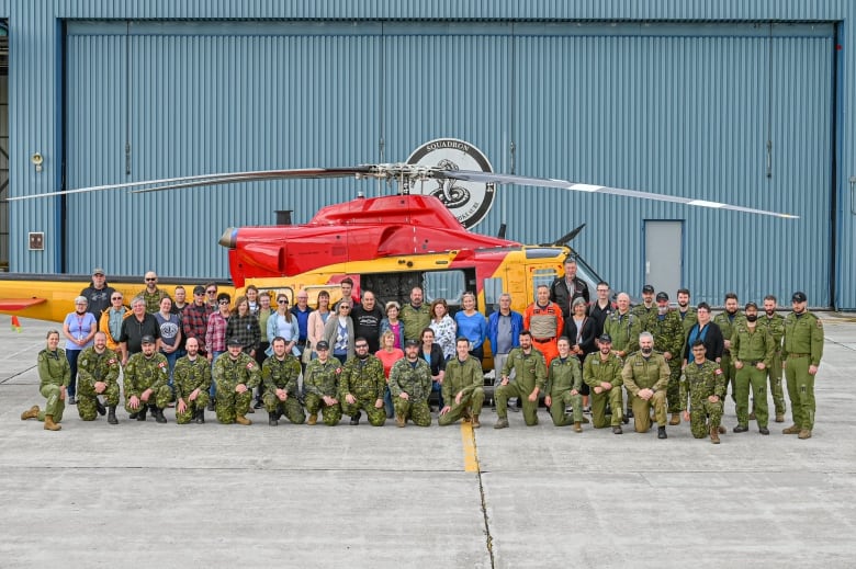 A group of people and military personnel stand in front of a yellow and red helicopter.