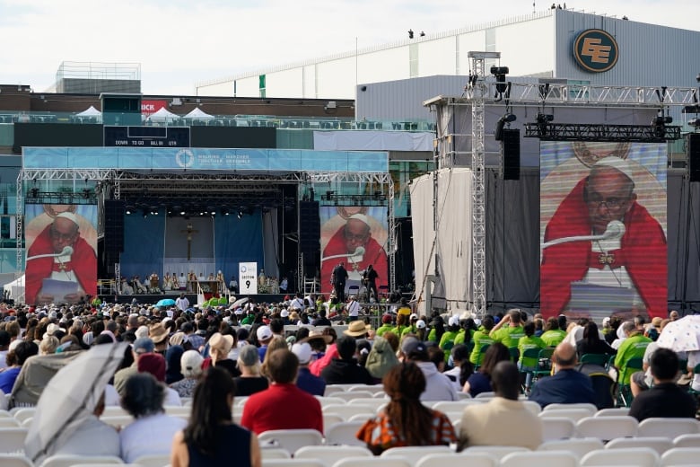 Crowds of people in an open air stadium look at giant screens showing Pope Francis