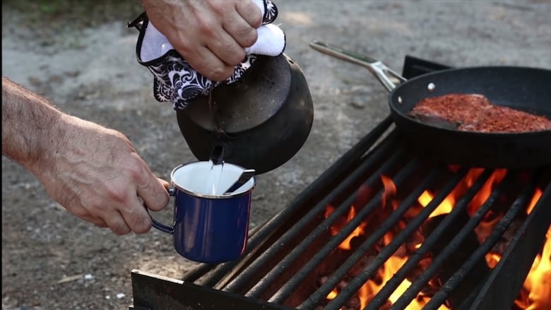 A person pours coffee from a coffee pot near a cooking campfire