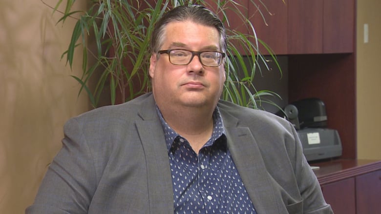 A man wearing glasses in a grey blazer over a blue and white shirt. 