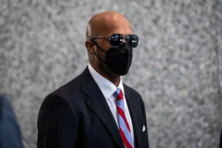 A man in a suit, dark sunglasses and a black mask is pictured in front of a grey wall.