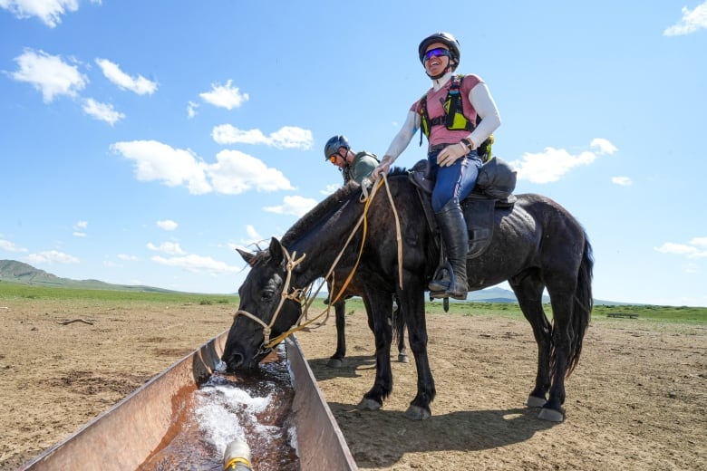 Two people on horses by a water trough. The nearest person is a woman and she is smiling. Her horse is drinking water. 