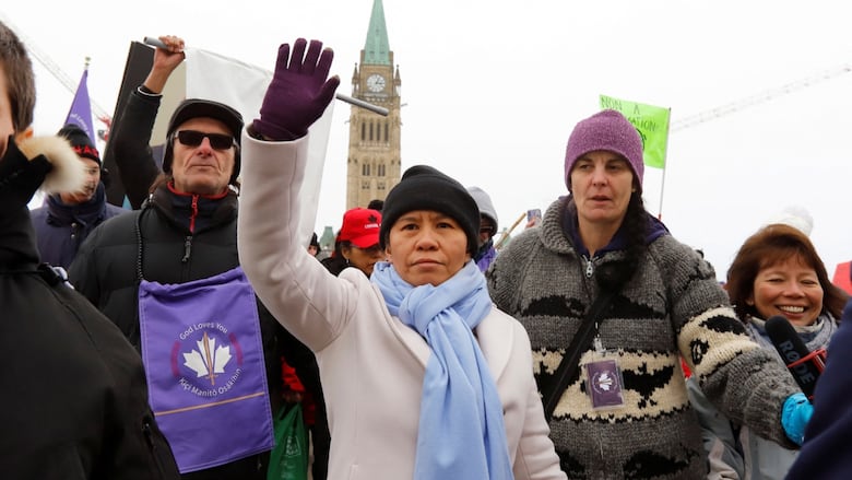 A crowd of people in winter clothes standing in front of Ottawa's Parliament building raise their hands in the air. The Parliament building's steeple with the clock and green tower are in the background.