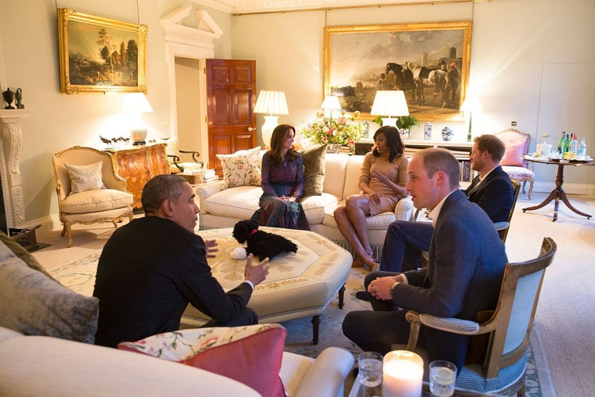 Five people talk among one another as they sit on couches and chairs in a large formal living room.