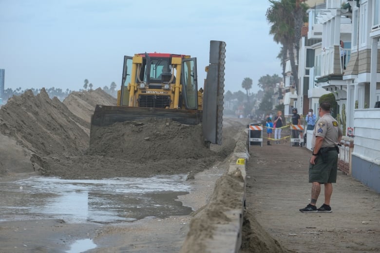 People stand in front of a row of houses as a bulldozer pushes sand past piles of sand.