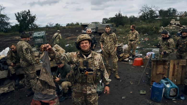 A Ukrainian soldier holds up the Russian flag, with his a bunch of comrades behind him in a countryside setting.