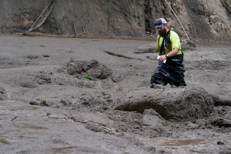 A man in a yellow t-shirt and black hip-waders pants walks through deep mud up to his knees.