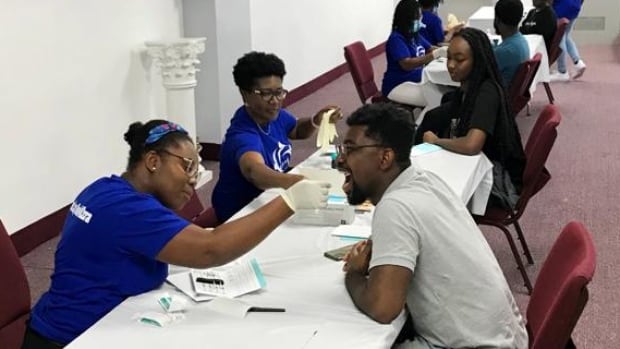 Stem-cell matching tests require a simple swabbing of the inside of the cheek, as volunteers at the Pentecost Church's Swab4Ezra event lined up to do on Sept 17. Members of the local Ghanian community are doing stem-cell matching tests, which involves swabbing the inside of their mouths. In front is one man getting his cheek swabbed by a volunteer medical personnel. Along the table are more volunteers and attendees to the event doing the same.
