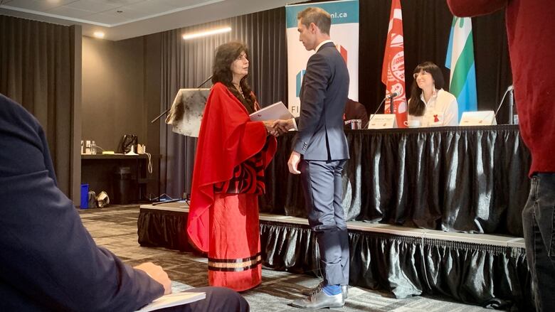 A woman in red hands a report to a man in a suit as people look on.