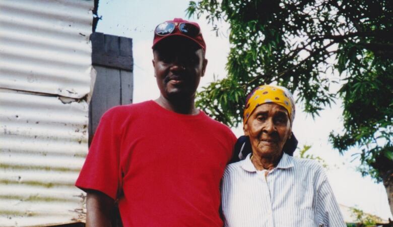 A man and older woman stand outside, in front of a tin-clad shack.