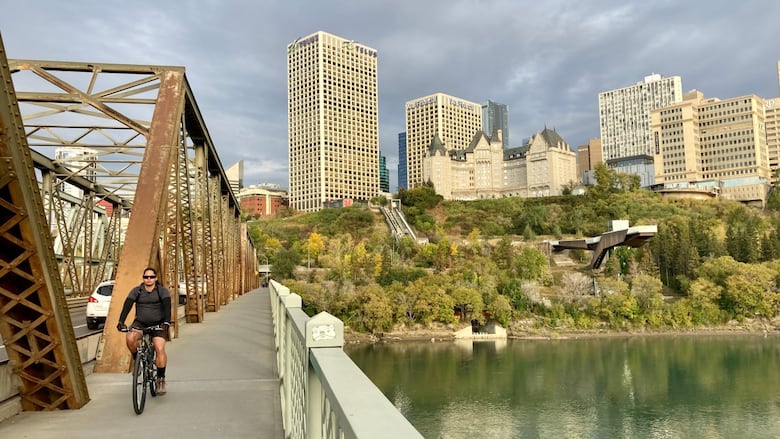A cyclist on a bridge over a river with city buildings in the background.