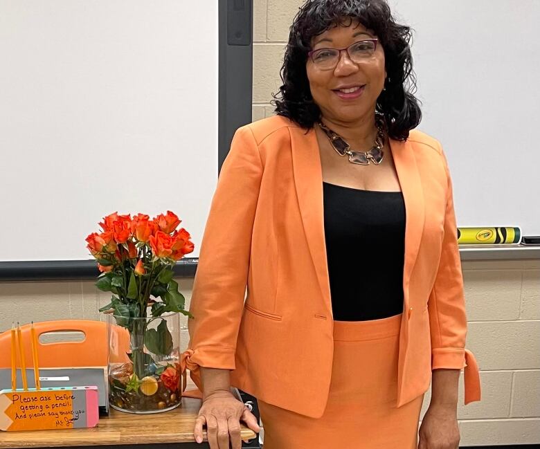 A teacher wearing a peach-coloured blazer and skirt stands at the front of a classroom next to her desk and a bouquet of orange roses in a vase.