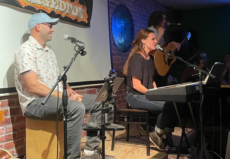 A three-piece band performs in a bar.  A man and a woman sit in front of a microphone while a second man stands in front of the microphone holding a guitar.