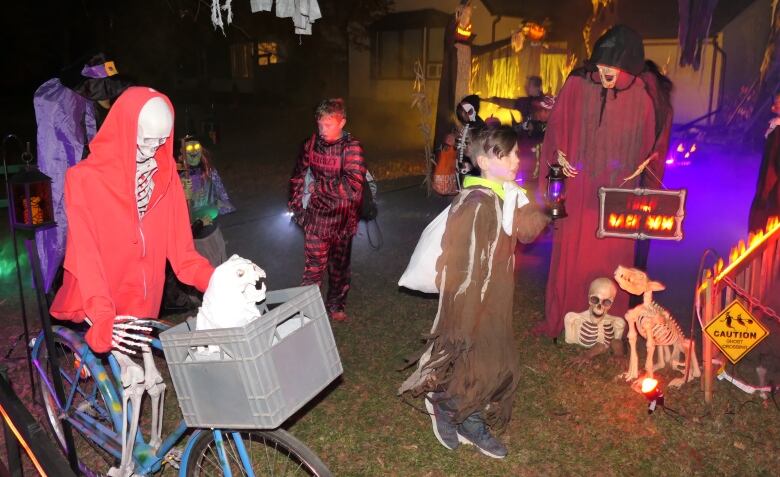 Two boys in Halloween costumes walk past several life-size Halloween figures, including a skeleton on a bike and a cloaked figure.