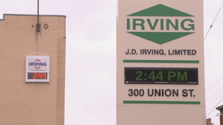 Two signs above two digital clocks are seen on nearby buildings. On the left, the red and blue logo of Irving Oil is seen. On the right is the sign for J.D. Irving, which simply says "Irving" in the colour green. The time on both clocks is 2:44;. 