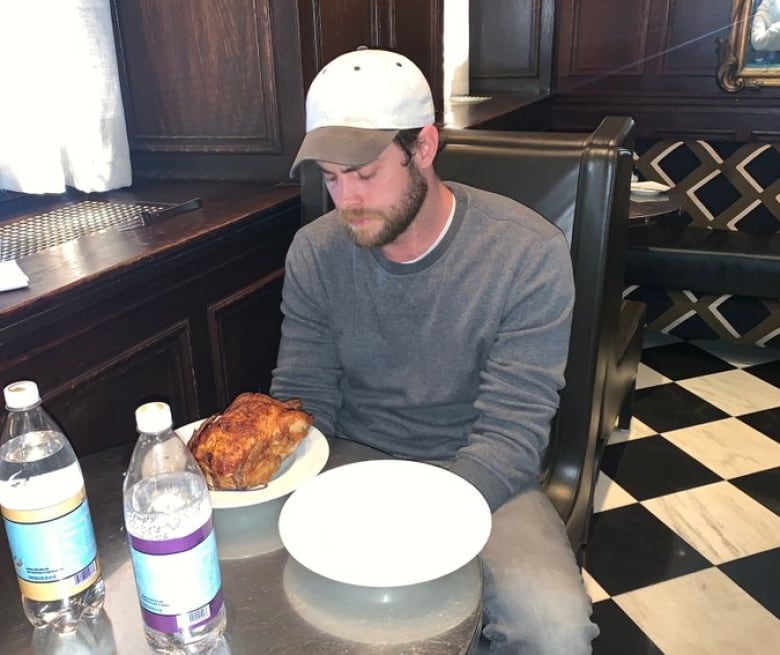 A man in a baseball cap sits at a table, looking forlornly at a rotisserie chicken on a plate.