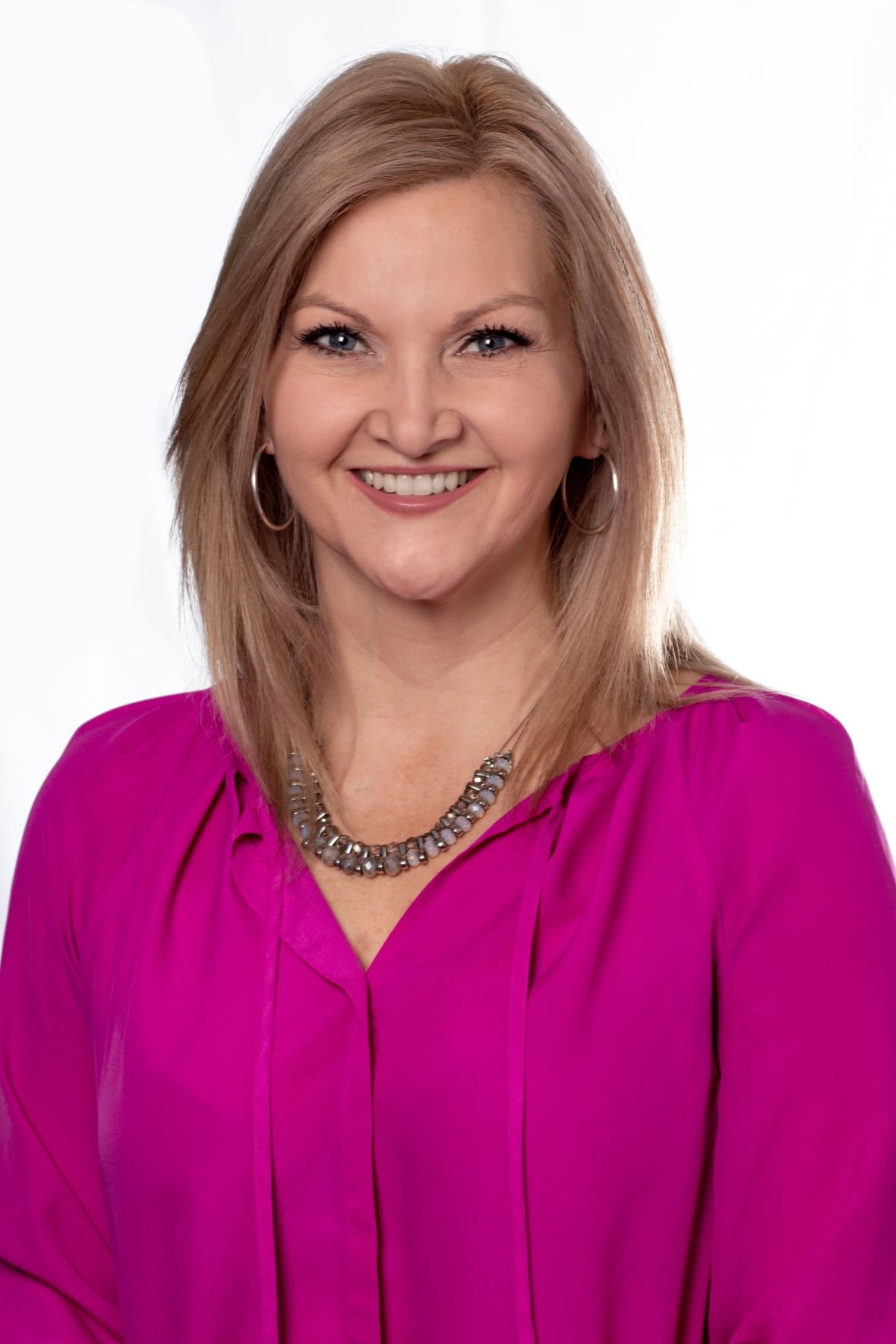 A head and shoulders professional portrait of a smiling woman with shoulder length straight blonde hair, parted on the side, wearing a fushia blouse, a fashion necklace that sits on the collarbone, and gold hoop earrings. 