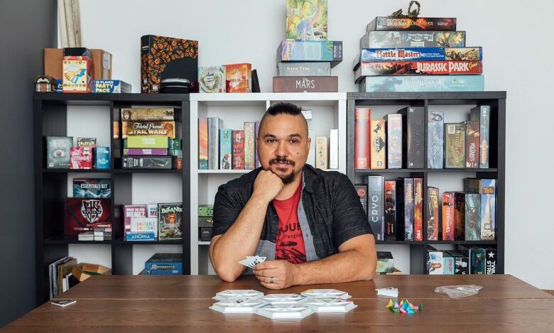 David Plamondon sits in front of shelves stuffed with board games, with one open on the table in front of him.