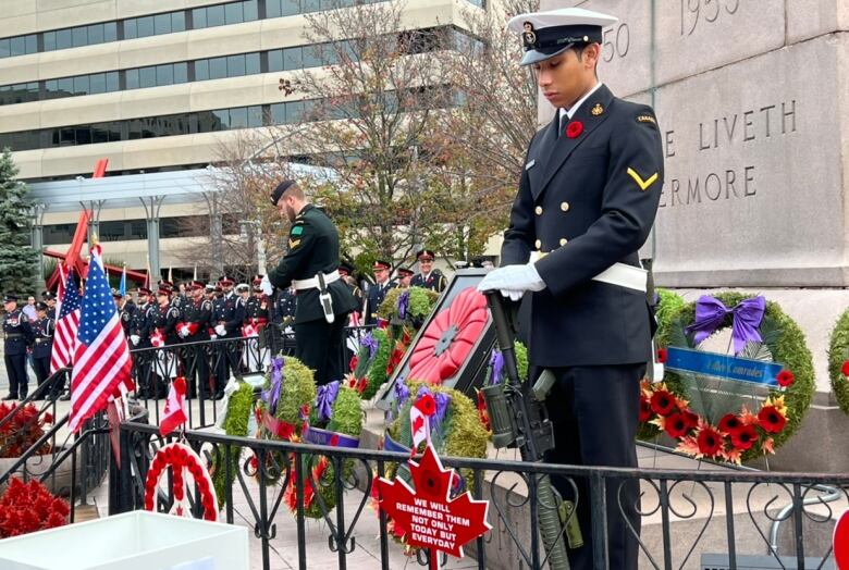 Two men in uniform stand in front of a giant cenotaph, surrounded by poppy-filled wreaths and flags.