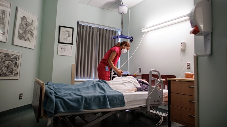 A woman in a red uniform leans over a hospital bed to tend to a person shrouded in sheets and blankets in what resembles a hospital room.