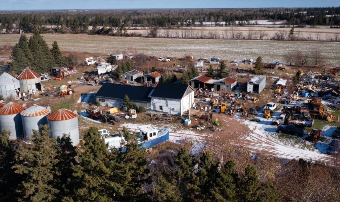 An aerial view of a farmhouse surrounded by rusted vehicles, boats on stakes, and small outbuildings. 