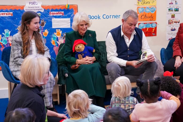 A person sits with a teddy bear on their lap as another person reads a story to children sitting in front of them.