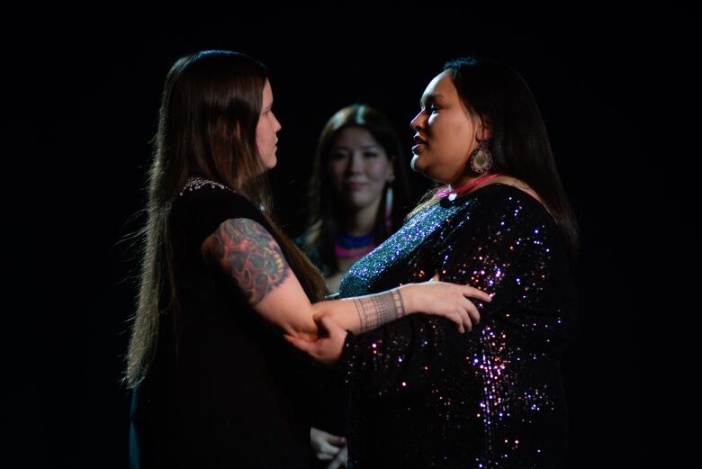 Three women stand in a close circle lit by stage lights. 