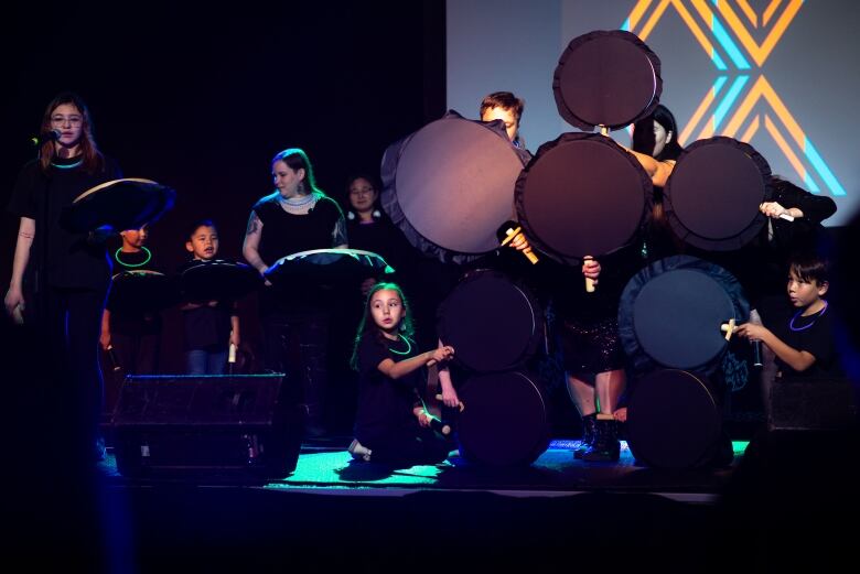 A group of children holding drums performing on a stage in a dimly lit room. 