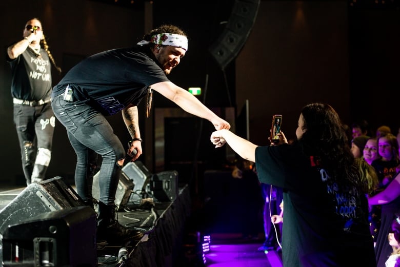 A man standing on a stage fist bumps a woman standing in a crowd. 
