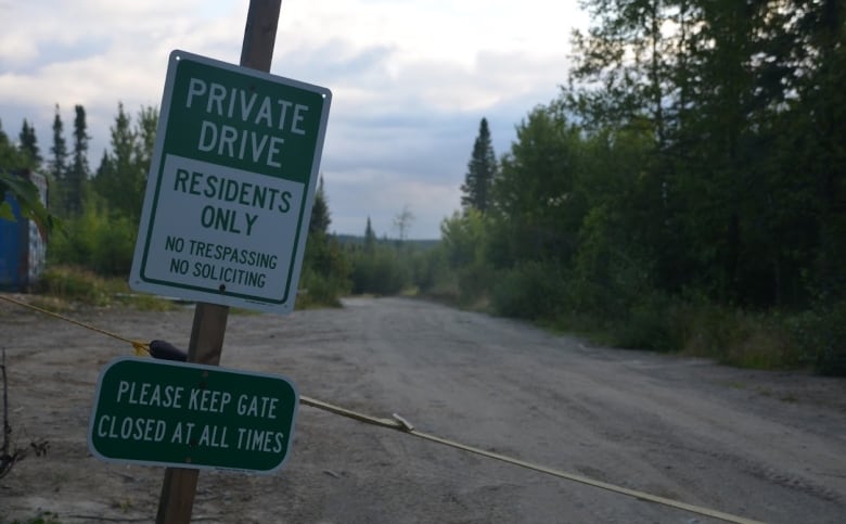 A dirt road leads into the wilderness past a small structure, a rope and a sign that reads 'Private drive: residents only'