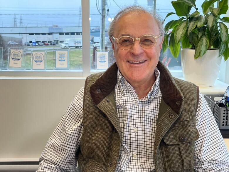 A smiling man in glasses, a checked dress shirt and checked vest sits at a desk, with a healthy green plant and a series of plaques displayed on the window sill behind him.