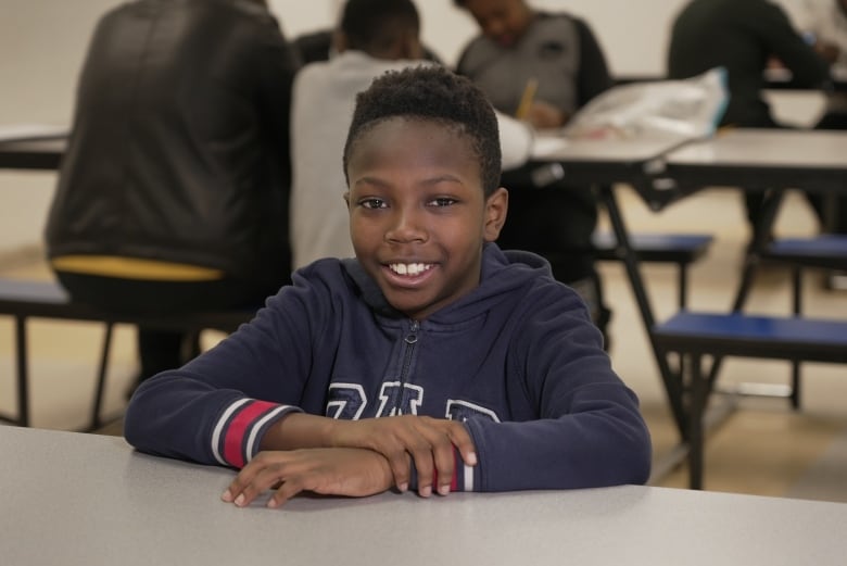 A young boy in a navy blue zip-up sweatshirt smiles while sitting at a table in a school space. Other students work with an adult tutor seated at tables and benches behind him.