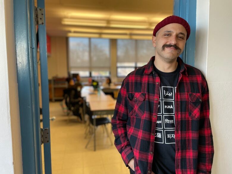 A mustachioed man wearing a knit cap and red and black plaid shirt over a black tee stands in the doorway, with students in a classroom seen out of focus behind him.