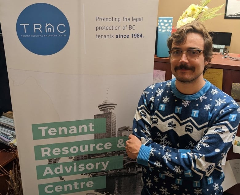 A man with glasses and a moustache stands in front of a banner for the Tenant Resource and Advisory Centre.