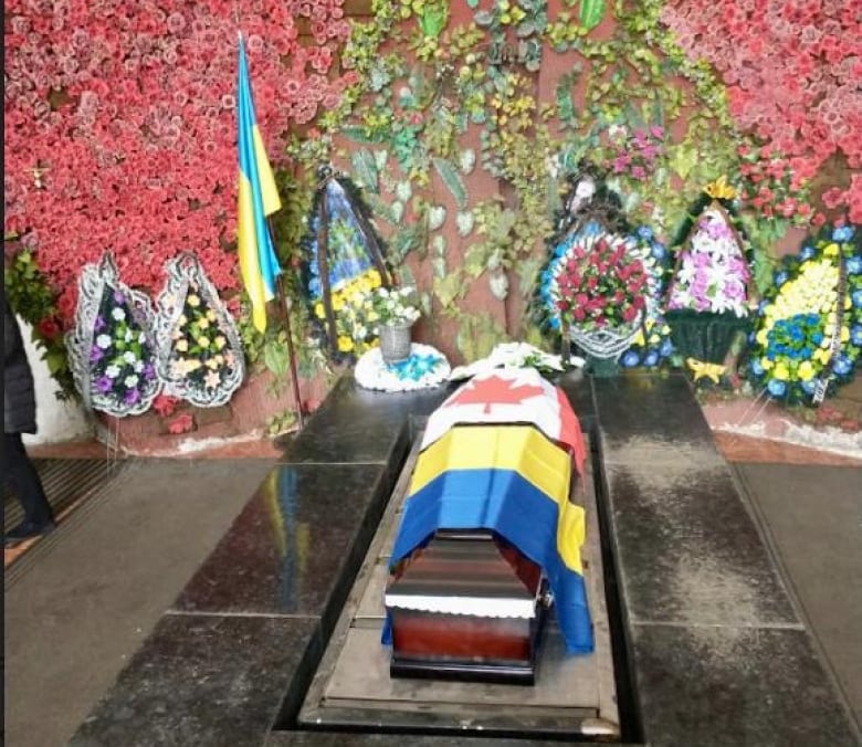 A casket with the Ukrainian and Canadian flags draped overtop sits in the middle of a room. There are flowers behind the casket.