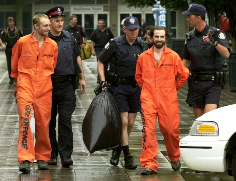 Two men wearing orange jumpsuits with the word 'Greenpeace' written on them are led away by three police officers.