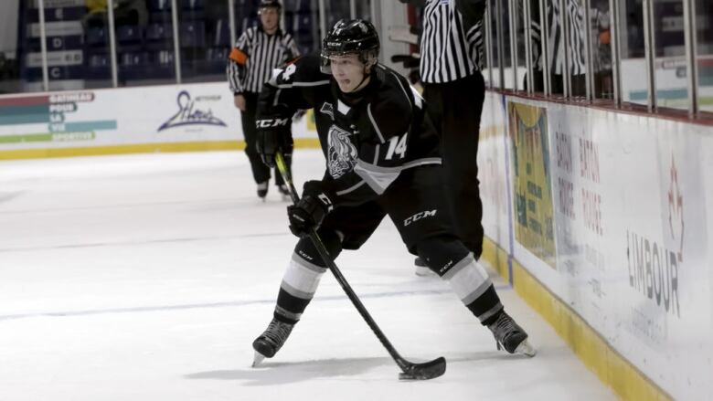 A hockey player wearing black. Two referees are in the background.