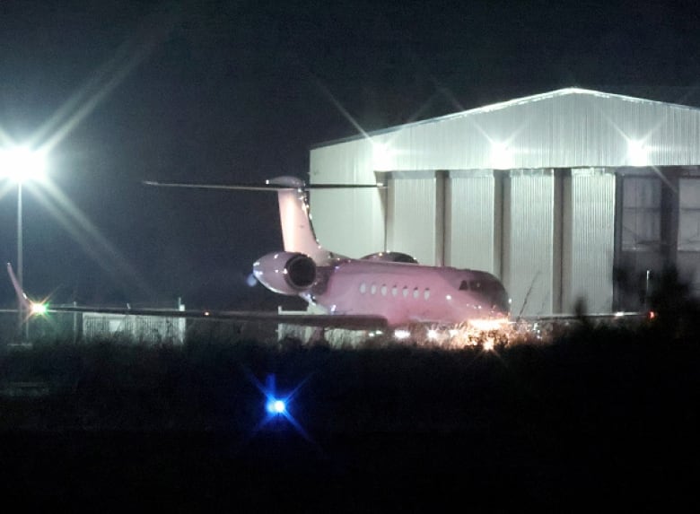 A white jet is seen on a runway at night with a green hangar in the background.