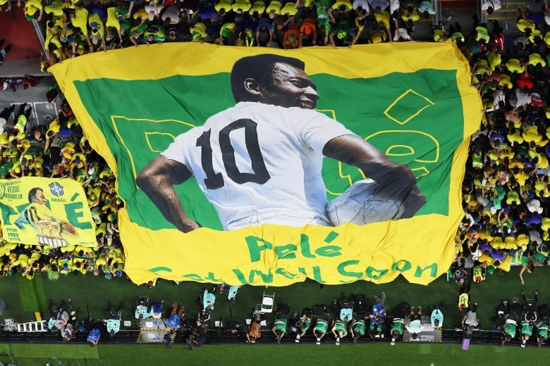 A banner reading "Pele, get well soon" is displayed by Brazil fans in the stands of a World Cup game. Pele is seen in the green and yellow flag in his playing days, holding the ball with his right hand. The number 10 is seen in his yellow shirt.