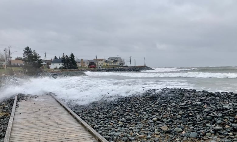 Waves wash over a wooden trail across rocks, while homes and trees are seen in the background.