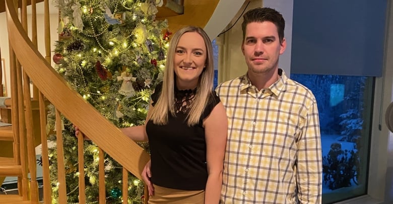 A man and woman stand next to a staircase with a decorated christmas tree behind them.