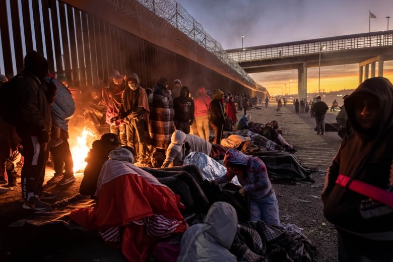 Several people are huddled near a bridge at dawn, some gathering around a campfire.