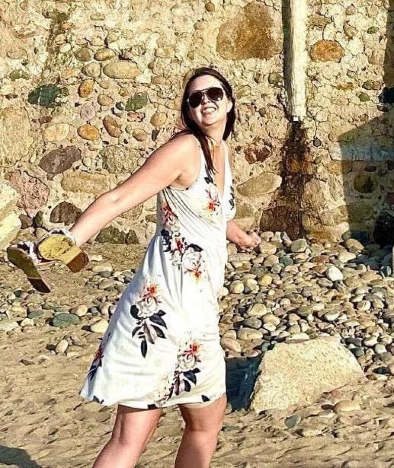 A young woman in a patterned white dress poses on a sunny beach.